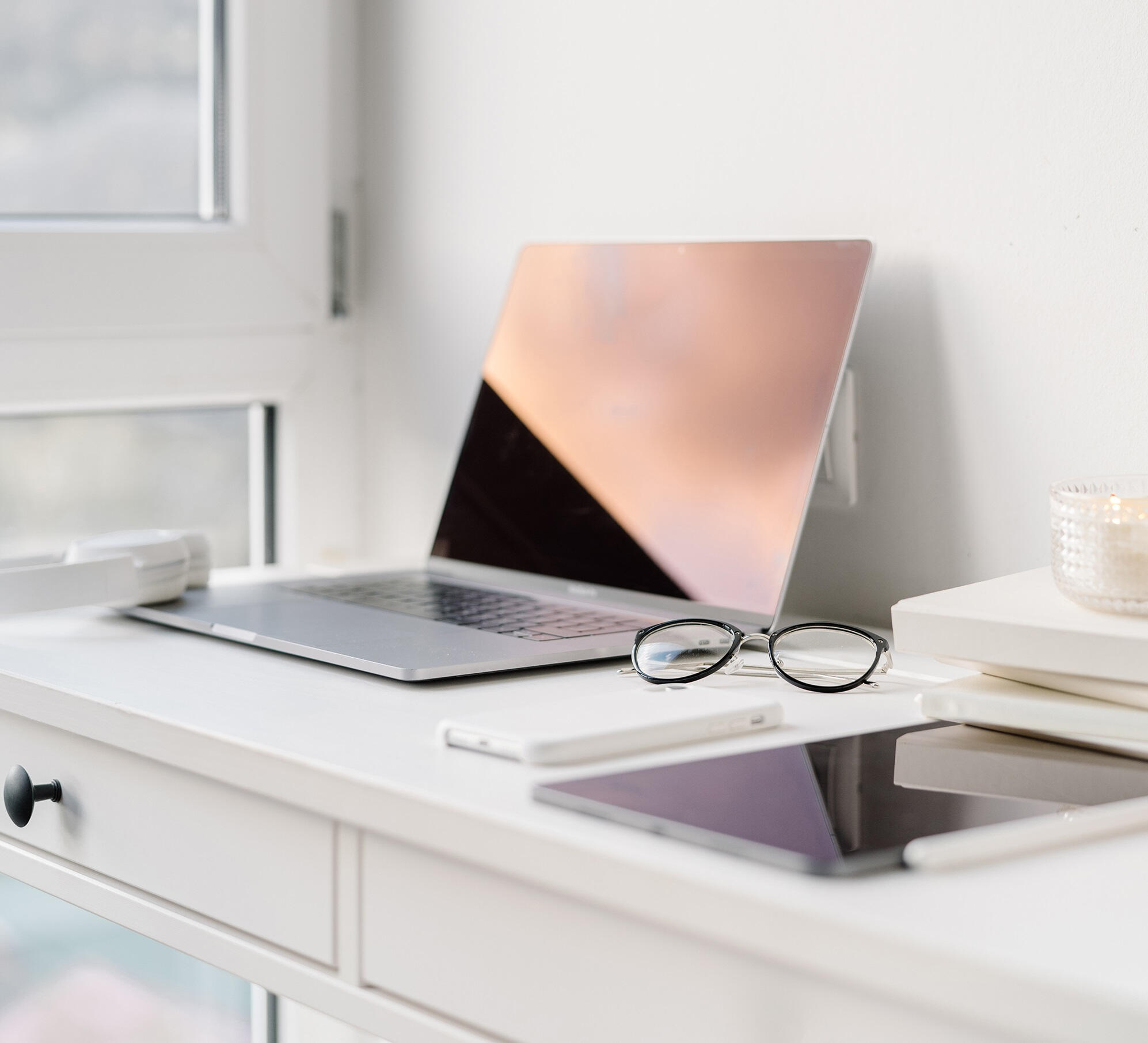 An open laptop, tablet computer, and reading glasses on a white desk next to a window.